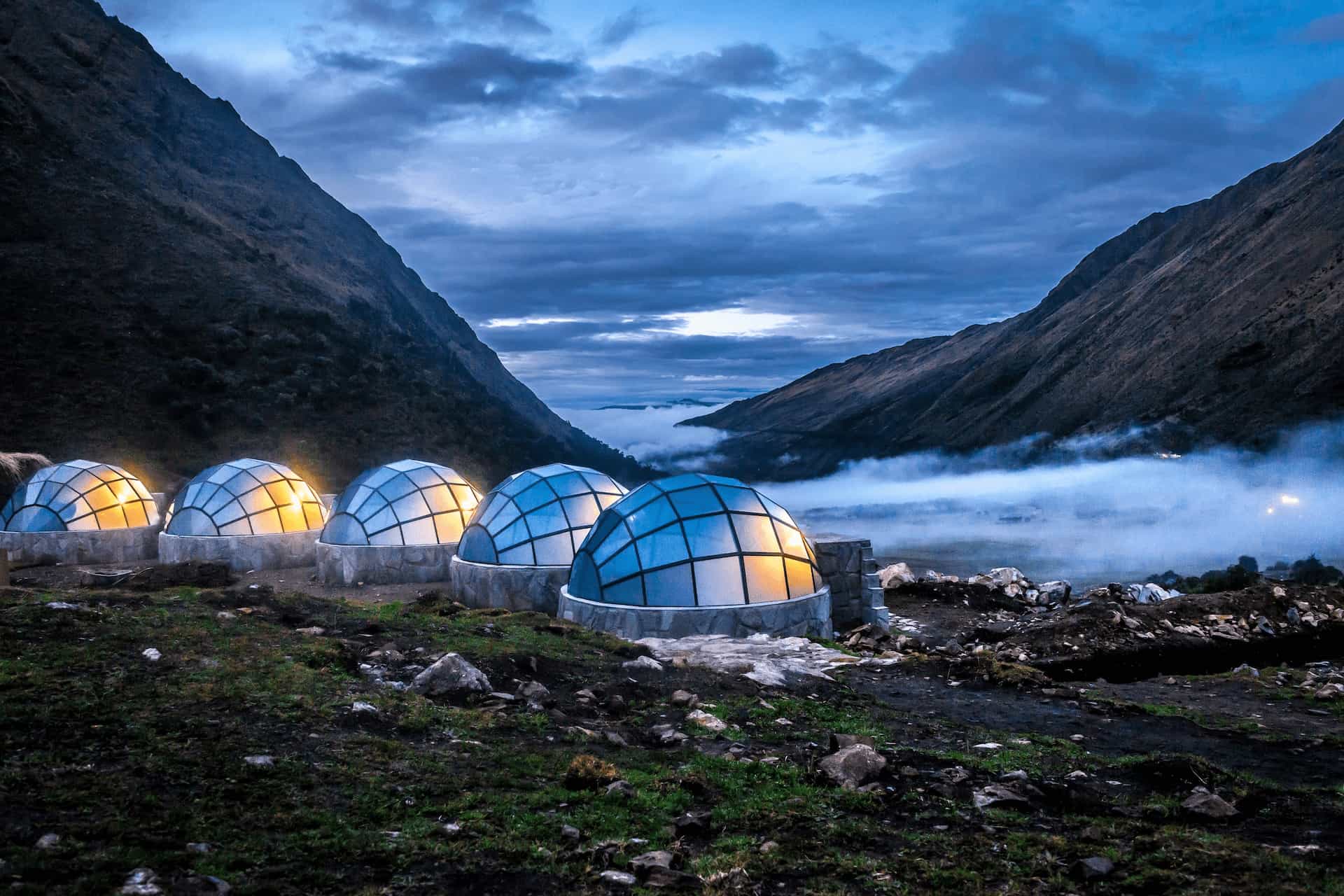 Glass-domed mountain camp glowing at dusk in a high Andean valley, cloud pooling below