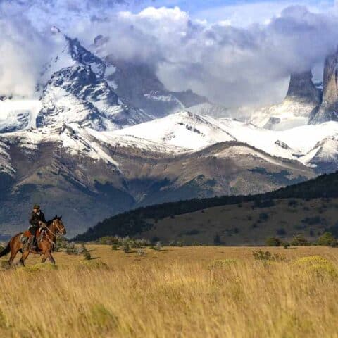 A rider on horseback crossing golden grassland below the snowbound Torres del Paine massif